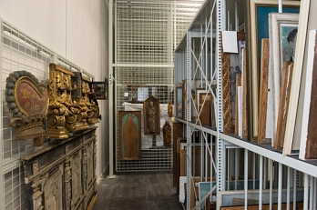 Digitale Depottour 2021 Foto aus dem Außendepot des Museums. Rechts stehen Gemälde in einem Regal. Links hängen Holzobjekte an der Wand. © Stadt Göttingen