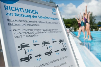 Foto aus einem Freibad am Brauweg. Rechts im Hintergrund spingen vier Personen im Badeanzug in das Schwimmbecken. Links im Vordergrund steht ein Schild auf dem  "Richtlinien zur Nutzung der Schwimmbecken"  steht. Unten auf dem Schild ist eine Grafik die schwimmende Personen darstellt. Zwischen den Personen sind Abstände von 2 oder 3 Meter eingezeichnet.&nbsp;&copy;&nbsp;Niklas Richter