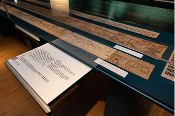 Foto aus der Sonderausstellung Gestickte Pracht und gemalte Welt. Zu sehen ist eine längliche Vitrine, in der drei Tora-Wimpel liegen. Auf einer herausgezogenen Schubladen steht Text.&nbsp;&copy;&nbsp;Stadt Göttingen