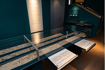 Foto aus der Sonderausstellung Gestickte Pracht und gemalte Welt. Zu sehen ist eine längliche Vitrine, in der drei Tora-Wimpel liegen. Auf zwei herausgezogenen Schubladen steht Text. An der Wand hinter der Vitrine hängt eine Textfahne.&nbsp;&copy;&nbsp;Stadt Göttingen